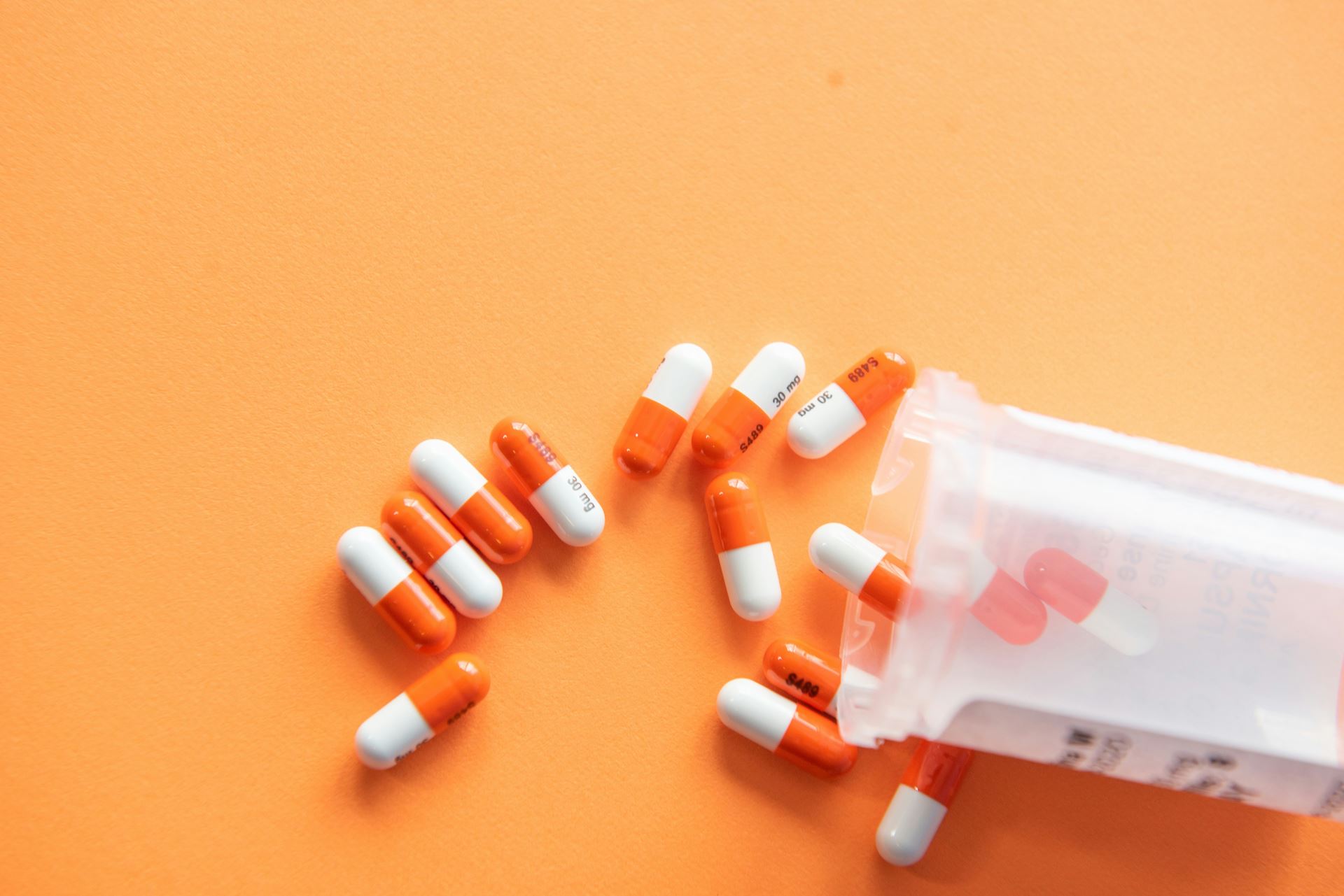 Orange and white medication capsules spilling out a container onto an orange background
